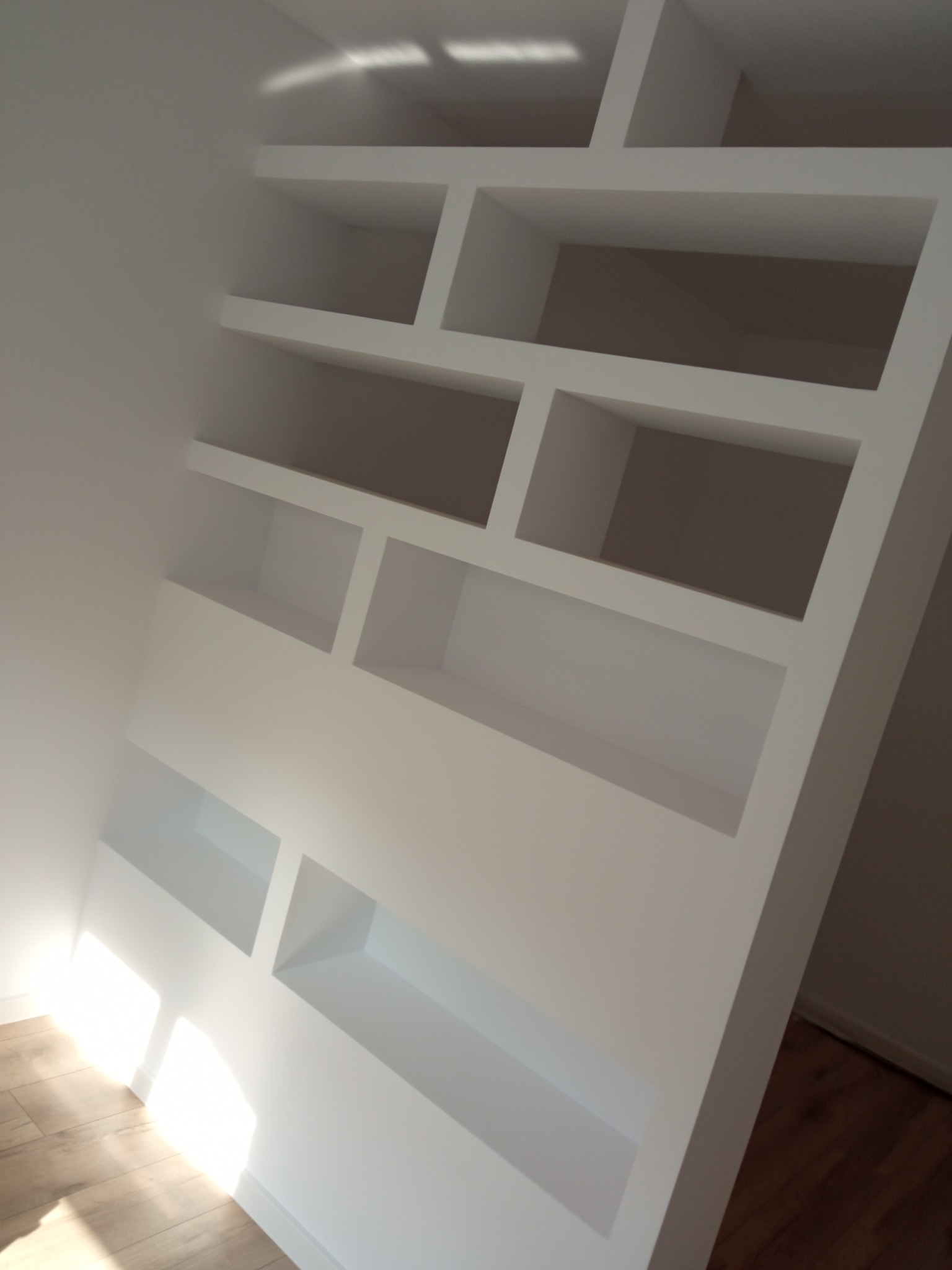 Wnęka ścienna z nieregularnymi, prostokątnymi półkami w kolorze białym, oświetlona naturalnym światłem padającym z góry, z widoczną częścią drewnianej podłogi.