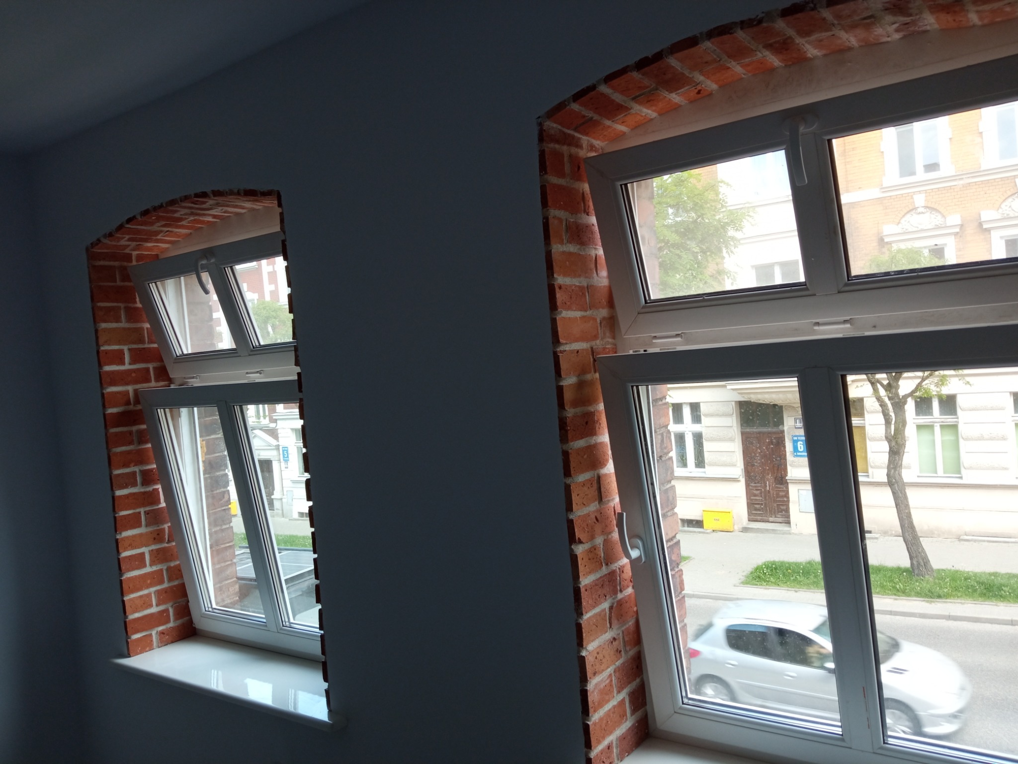 Dwa okna z białymi ramami i ceglanym obramowaniem w nowo wykończonym wnętrzu, widok na ulicę z przejeżdżającym samochodem.