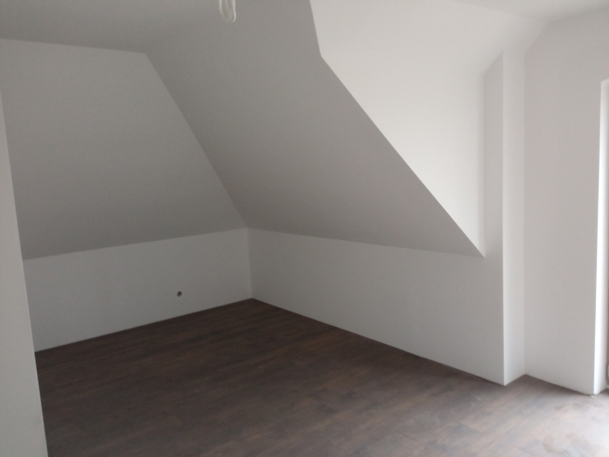 Poddasze z wykończonymi ścianami i ciemną podłogą z paneli, widoczne skosy i narożniki pomieszczenia.