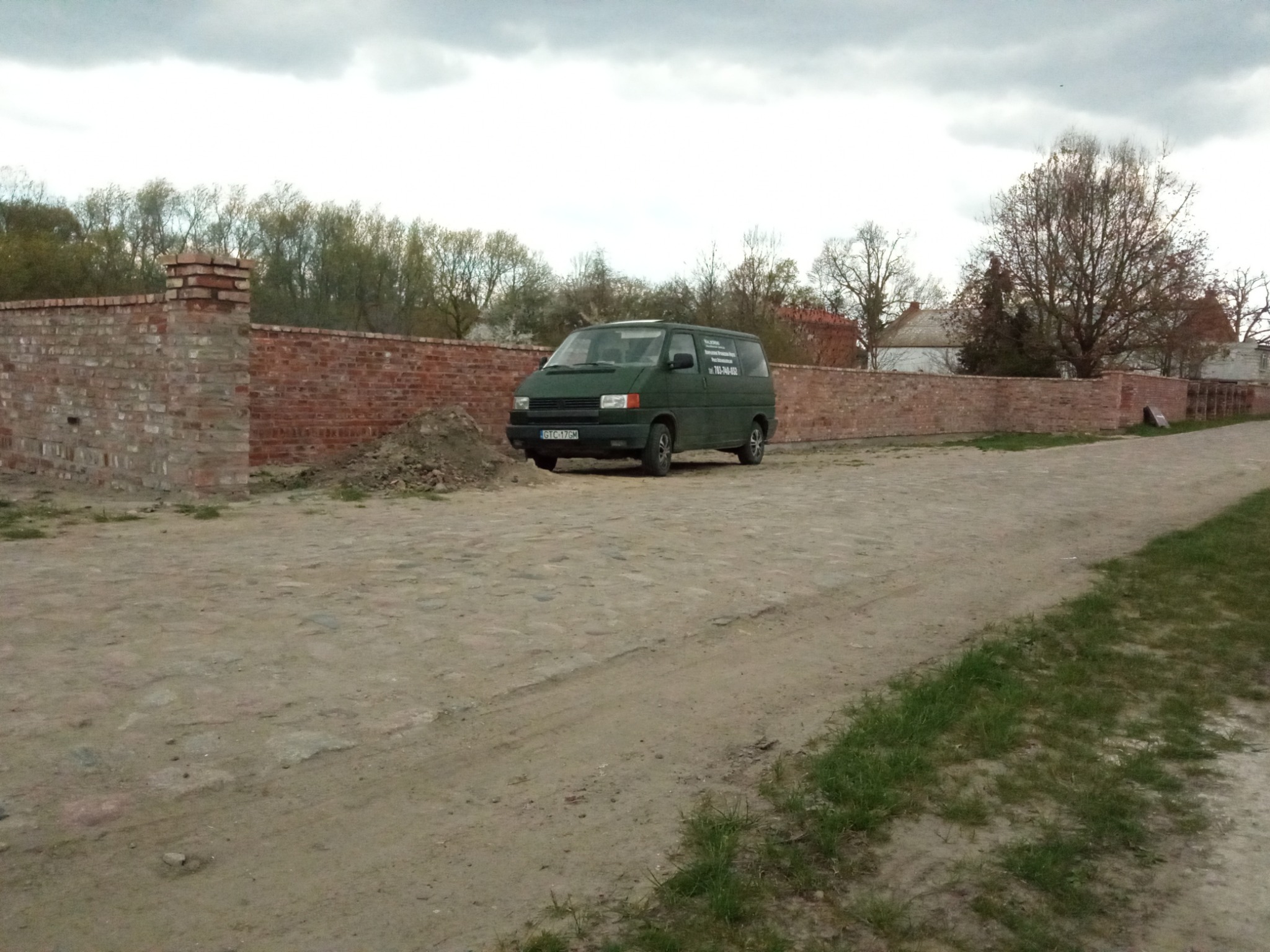 Zielony bus firmy budowlanej zaparkowany na brukowanej drodze przed murem z czerwonej cegły, w tle drzewa i pochmurne niebo.