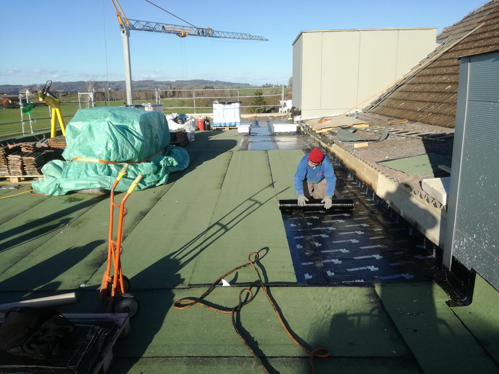 Pracownik w czerwonej czapce układa czarny pas hydroizolacji na zielonym dachu płaskim, w tle widoczne materiały budowlane, palety z dachówkami i żuraw budowlany.