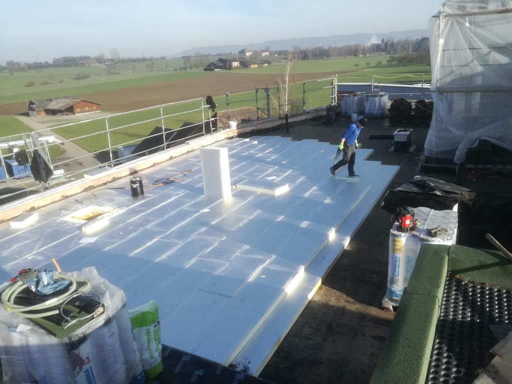 Układanie białych płyt izolacyjnych na płaskim dachu, widoczny pracownik w niebieskiej bluzie, butla z gazem, narzędzia i krajobraz w tle.