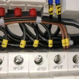 Szczegółowe ujęcie podłączonych przewodów elektrycznych w skrzynce rozdzielczej, z oznacznikami na kablach i widocznymi modelami bezpieczników.