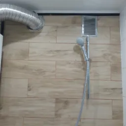 Bojler z aluminiową rurą odprowadzającą spaliny, zamontowany na ścianie wyłożonej płytkami imitującymi drewno, obok widoczny panel prysznicowy z deszczownicą i kratka wentylacyjna.