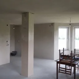 Surowe wnętrze w trakcie prac wykończeniowych: ściany pokryte świeżo nałożoną warstwą, widoczne niedoskonałości na filarach, prowizoryczne oświetlenie z wiszącymi żarówkami, stare krzesła i stół...