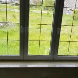 Nowe, białe okno trzyskrzydłowe z widokiem na zielone pole, parapet wykończony jasnymi płytkami, surowe ściany w tle przed malowaniem.