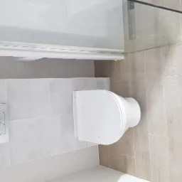 Nowoczesna łazienka po remoncie: podwieszana toaleta z chromowanym przyciskiem spłukującym, beżowe płytki imitujące drewno na podłodze i fragment szklanej kabiny prysznicowej.