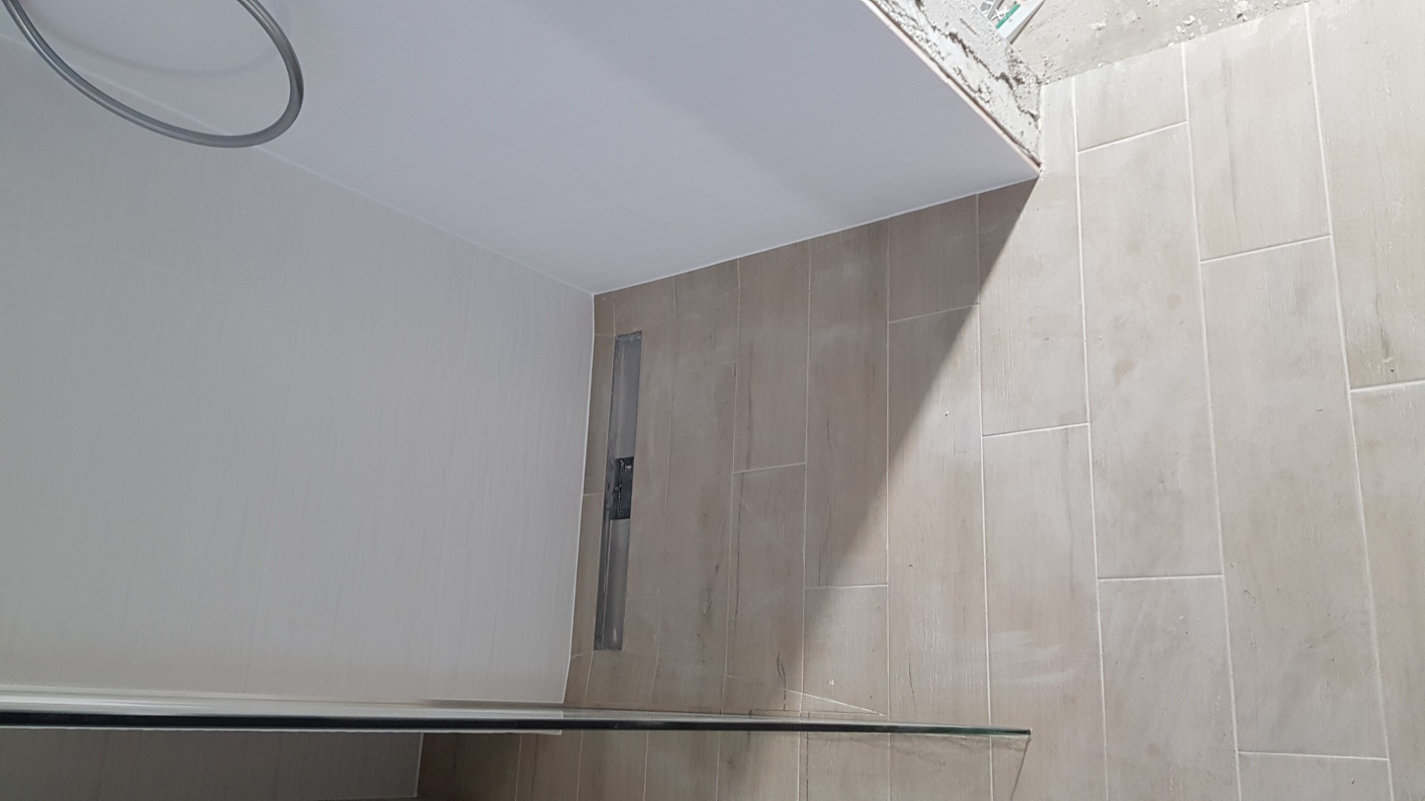 Świeżo ułożone, pionowe płytki w beżowym odcieniu na ścianie łazienki, z widocznym odpływem liniowym i fragmentem sufitu podwieszanego.