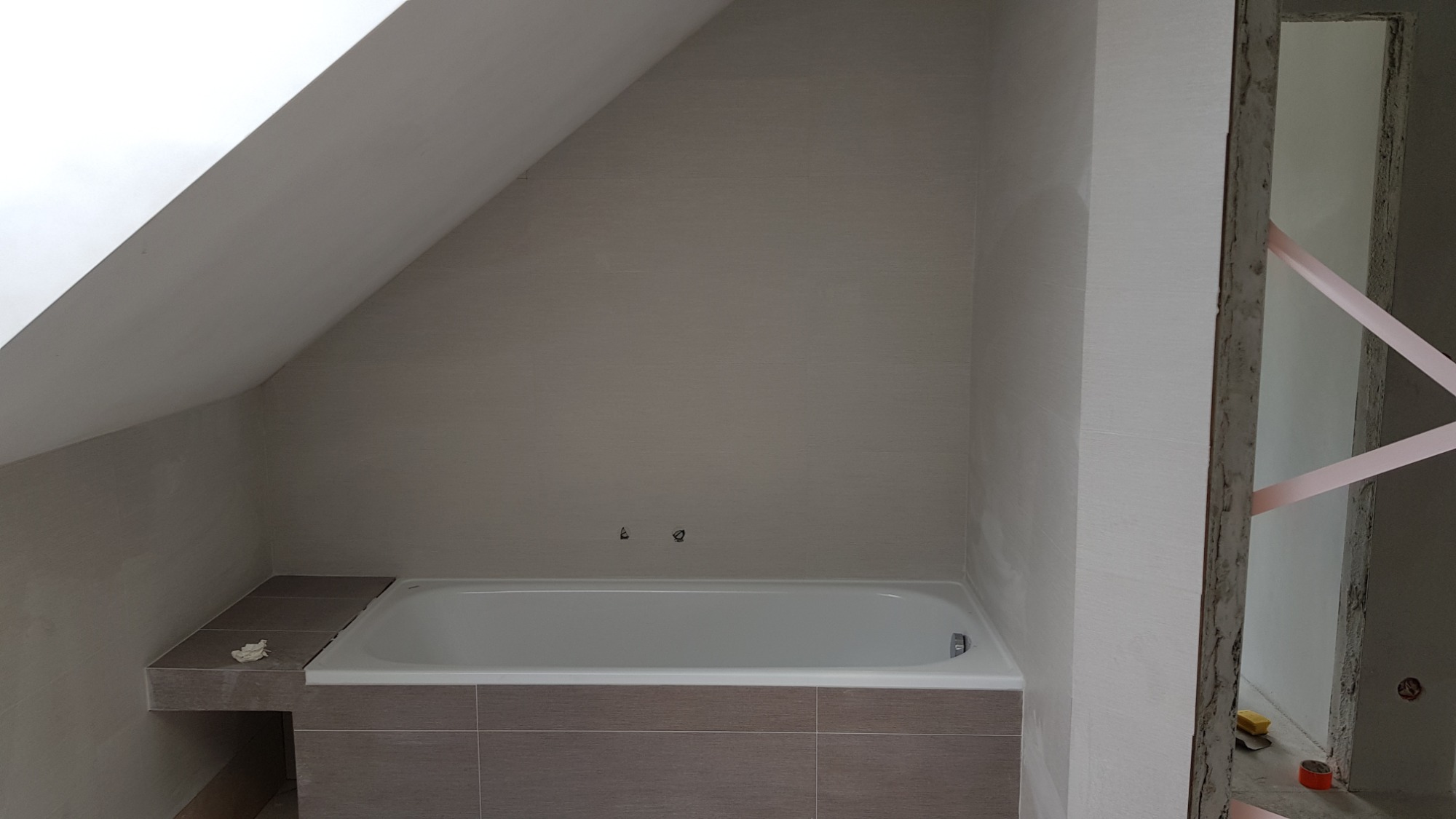 Niedokończona łazienka na poddaszu z wanną obudowaną brązowymi płytkami, widoczne surowe ściany przygotowane do malowania, fragment otworu drzwiowego zabezpieczonego taśmą.