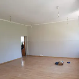 Pomalowane na szaro ściany i ułożona podłoga z paneli w pustym pomieszczeniu z widocznymi śladami po remoncie, na podłodze leżą ubrania i papierowa torba.