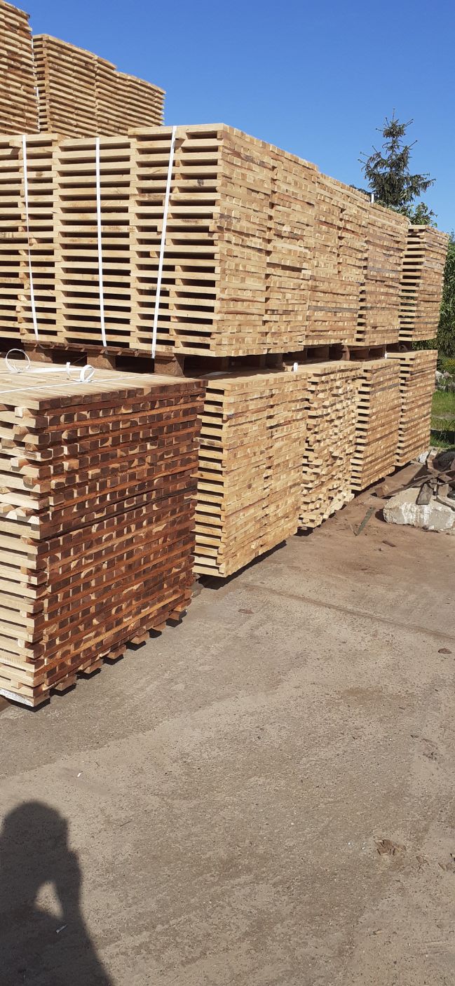 Stosy świeżo przetartego drewna ułożone na paletach, zabezpieczone białymi taśmami, na tle betonowego placu i zieleni.