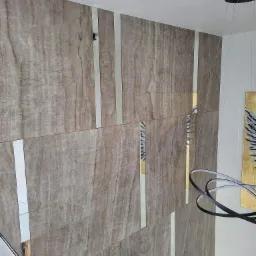 Ściana wykończona panelami imitującymi marmur, z wstawkami z wąskich luster, oświetlona nowoczesną lampą wiszącą z czarnymi obręczami.