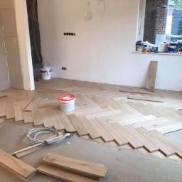 Usługi remontowe Damian Ujejski - Układanie jasnego parkietu w jodełkę francuską w trakcie prac wykończeniowych. Widoczne kleje, narzędzia i nieukończona powierzchnia podłogi.