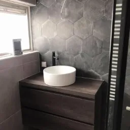 Nowoczesna łazienka z umywalką nablatową, heksagonalnymi płytkami w odcieniach szarości i szafką z ciemnego drewna. Widoczny fragment okna z roletą i niezakończone podłączenie elektryczne.