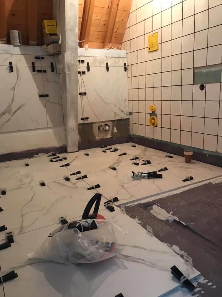 Łazienka w trakcie kładzenia płytek imitujących marmur na podłodze i częściowo na ścianach, z widocznymi krzyżykami dystansowymi i narzędziami, surowy strop z drewnianych belek.