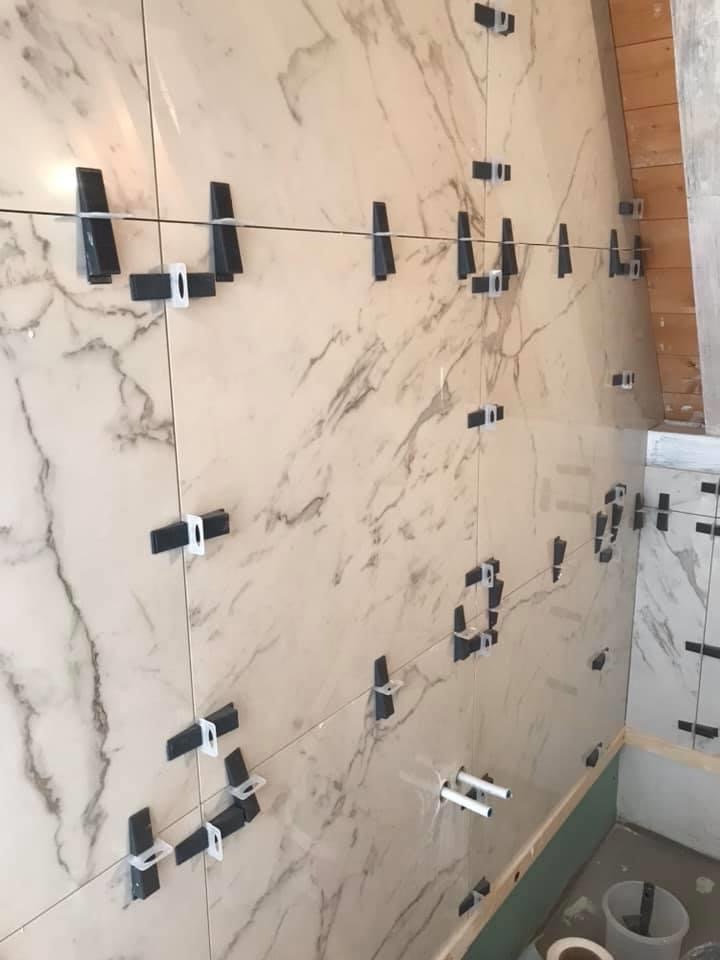 Ściana z marmurowych płytek, w trakcie montażu, z użyciem systemu poziomowania płytek (klipsy i kliny), widoczne rury instalacji wodnej i wiadro na podłodze.
