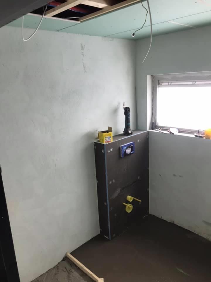 Surowa łazienka w trakcie prac wykończeniowych: widoczne stelaż podtynkowy WC z wystającymi rurami, częściowo wykończone ściany, sufit z odsłoniętą konstrukcją i wystającymi kablami, narzędzie...