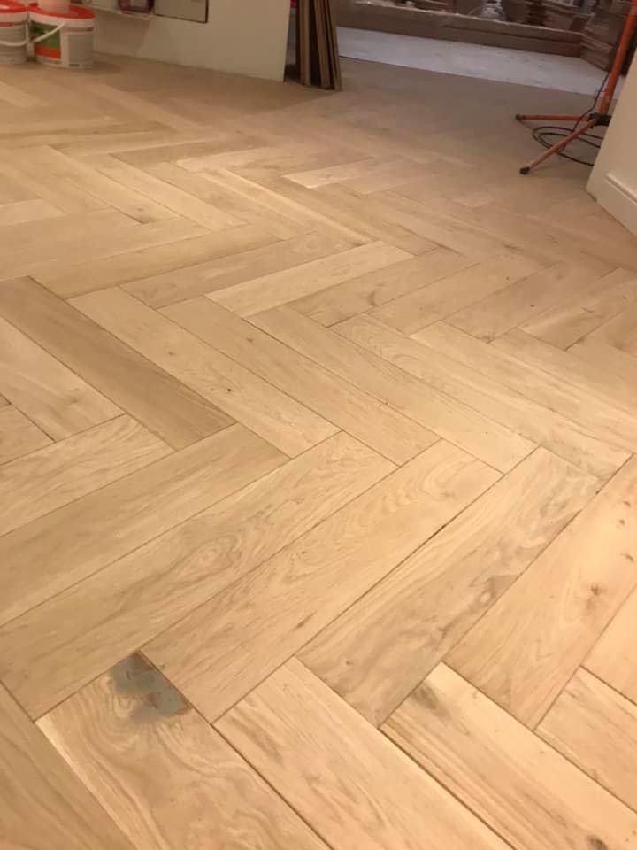 Jasna drewniana podłoga ułożona w jodełkę, widoczne wiaderka z farbą i narzędzia w tle.