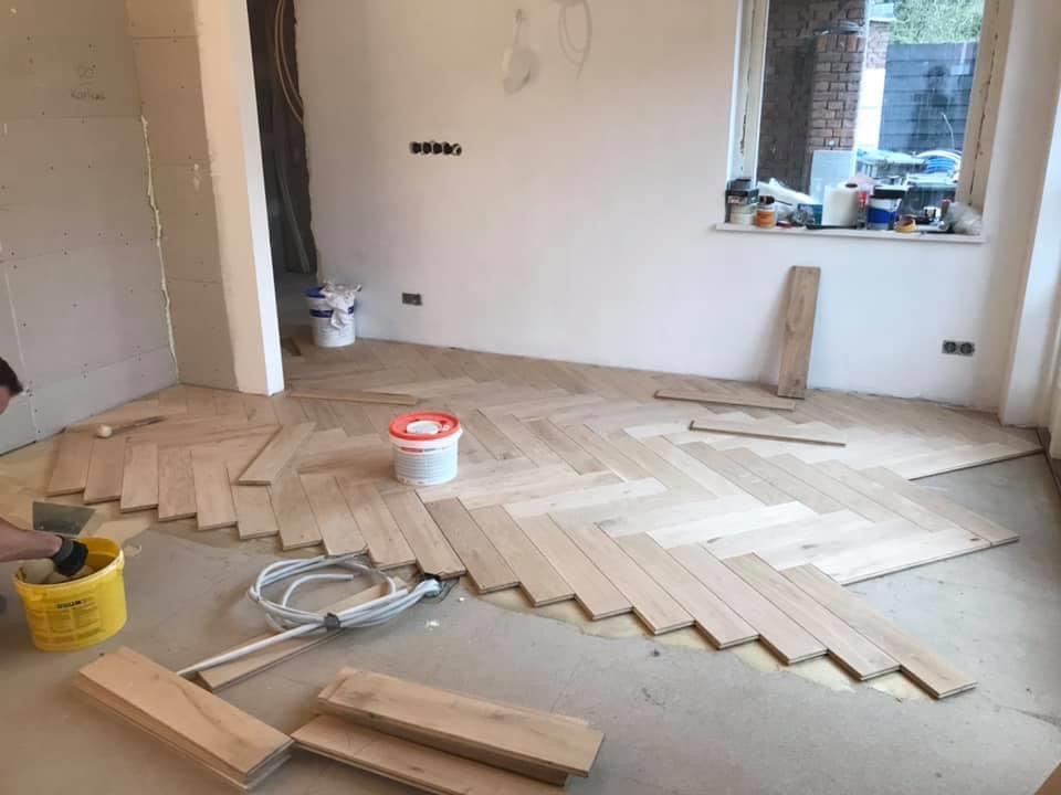 Układanie jasnego parkietu w jodełkę francuską w trakcie prac wykończeniowych. Widoczne kleje, narzędzia i nieukończona powierzchnia podłogi.