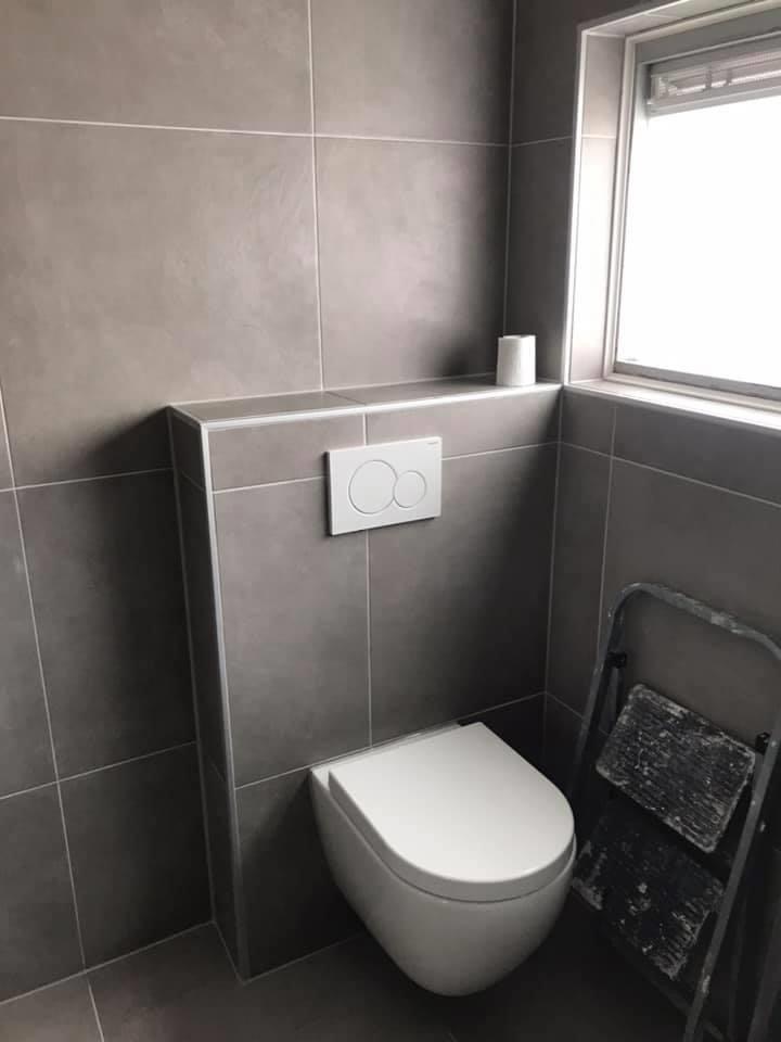 Nowoczesna toaleta podwieszana w szarej, wykafelkowanej łazience z drabinką opartą o ścianę i rolką papieru toaletowego na półce.