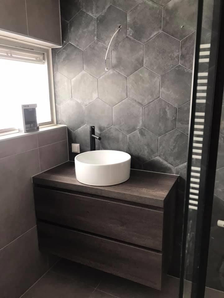Nowoczesna łazienka z umywalką nablatową, heksagonalnymi płytkami w odcieniach szarości i szafką z ciemnego drewna. Widoczny fragment okna z roletą i niezakończone podłączenie elektryczne.
