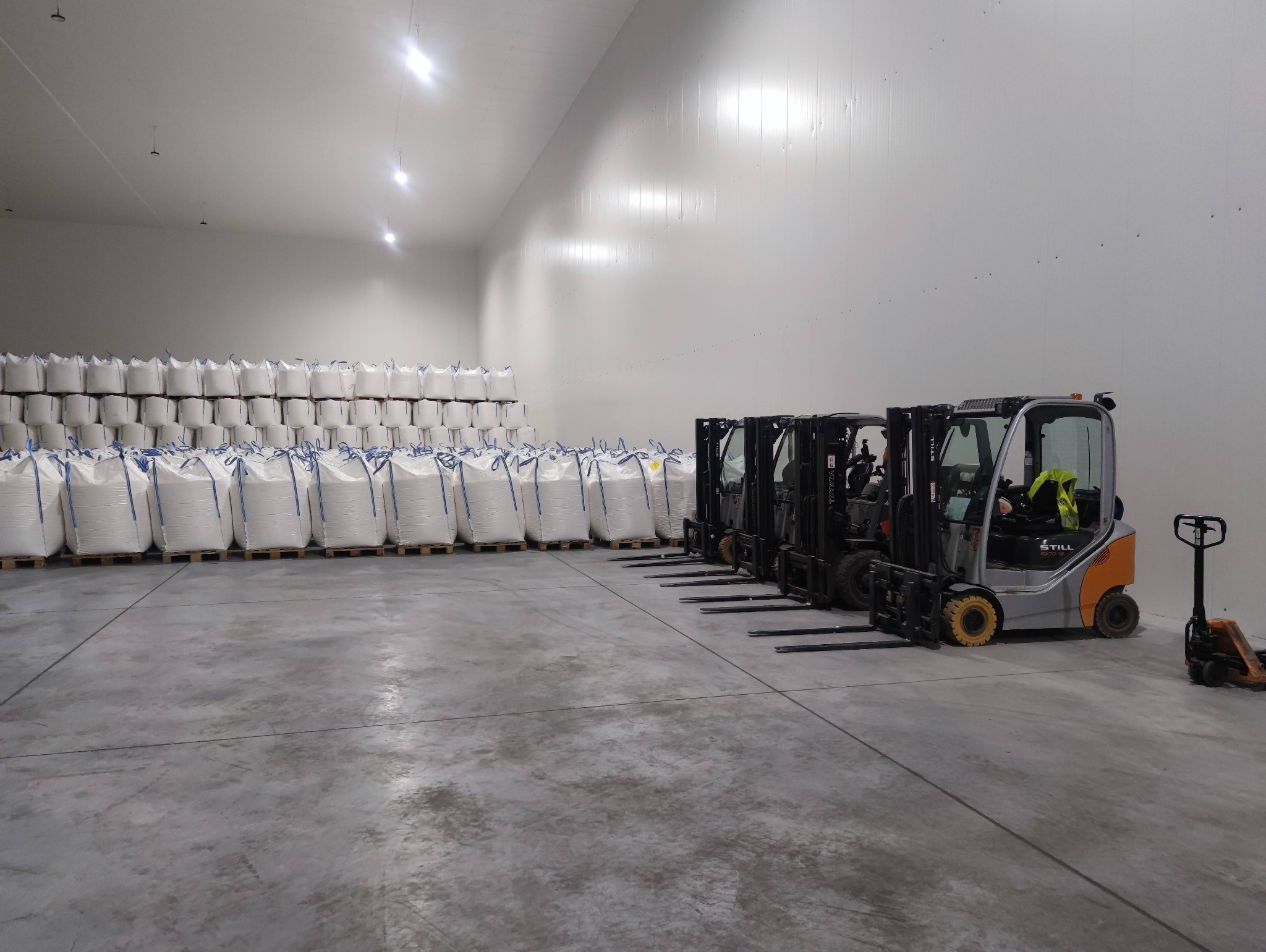 Wnętrze magazynu z rzędami palet z białymi workami i zaparkowanymi wózkami widłowymi STILL, oświetlenie sufitowe