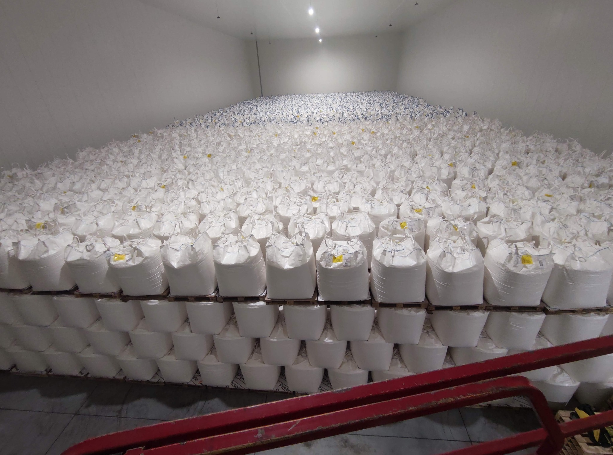 Wnętrze magazynu wypełnione paletami z białymi workami typu Big Bag, widoczne czerwone elementy wózka paletowego na pierwszym planie.