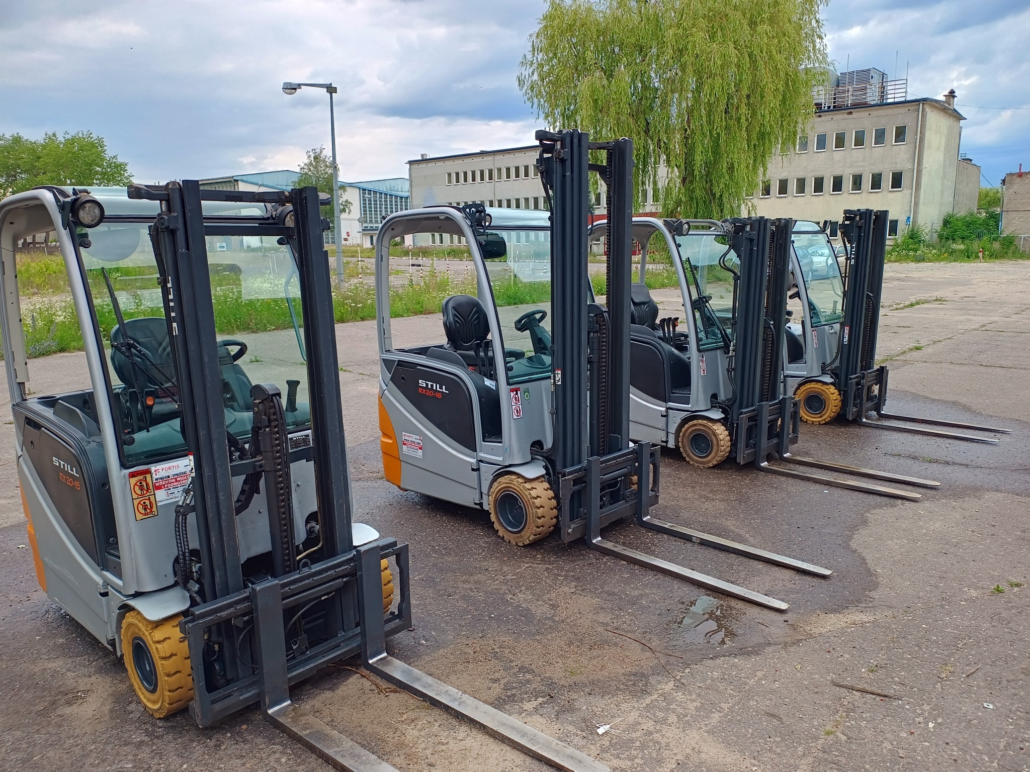 Rząd czterech wózków widłowych STILL RX20-15 zaparkowanych na betonowym placu przed budynkiem magazynowym, z widocznymi żółtymi oponami i czarnymi masztami