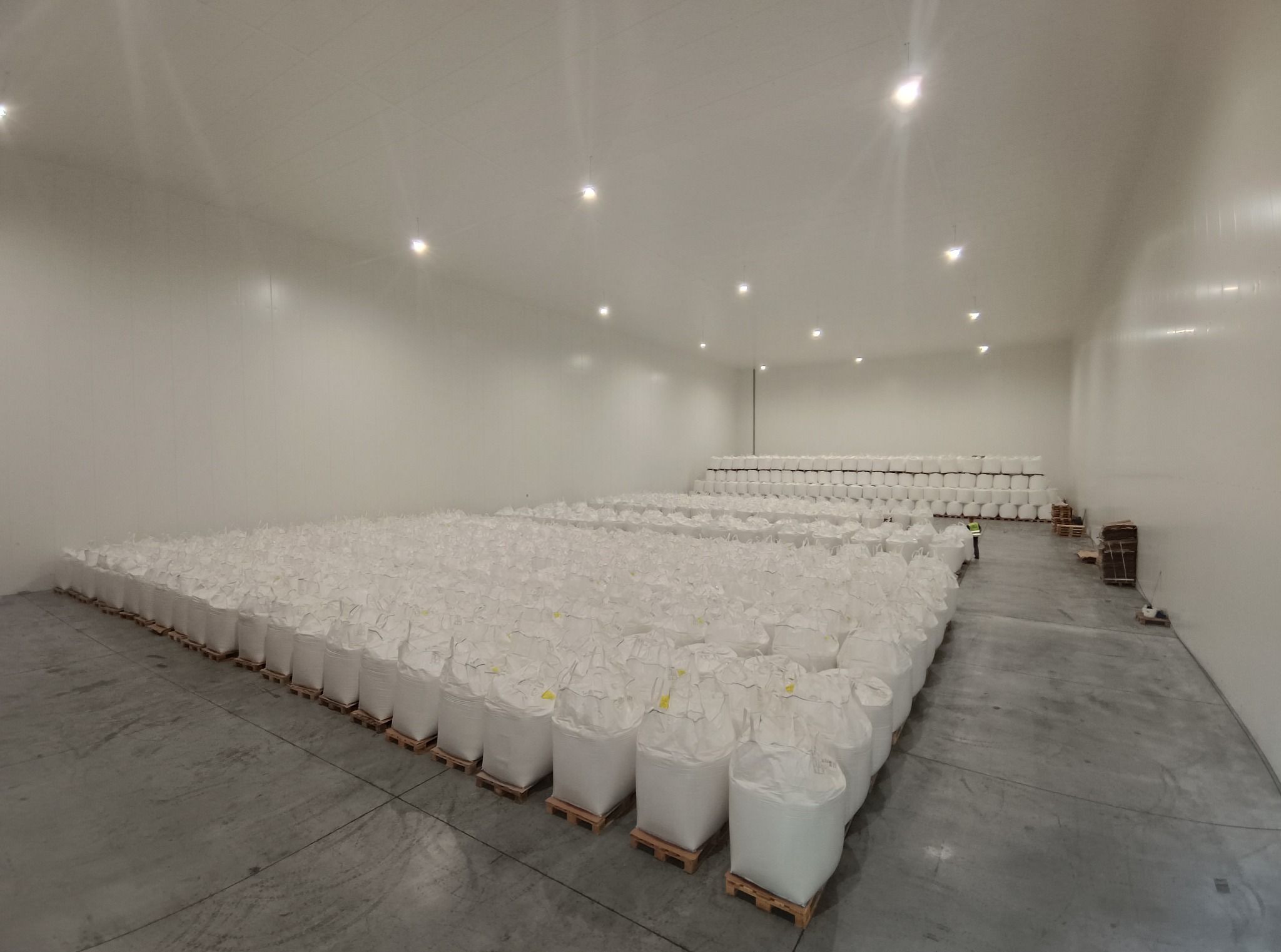 Wnętrze magazynu z rzędami ułożonych, białych worków na drewnianych paletach pod jasnym oświetleniem LED.