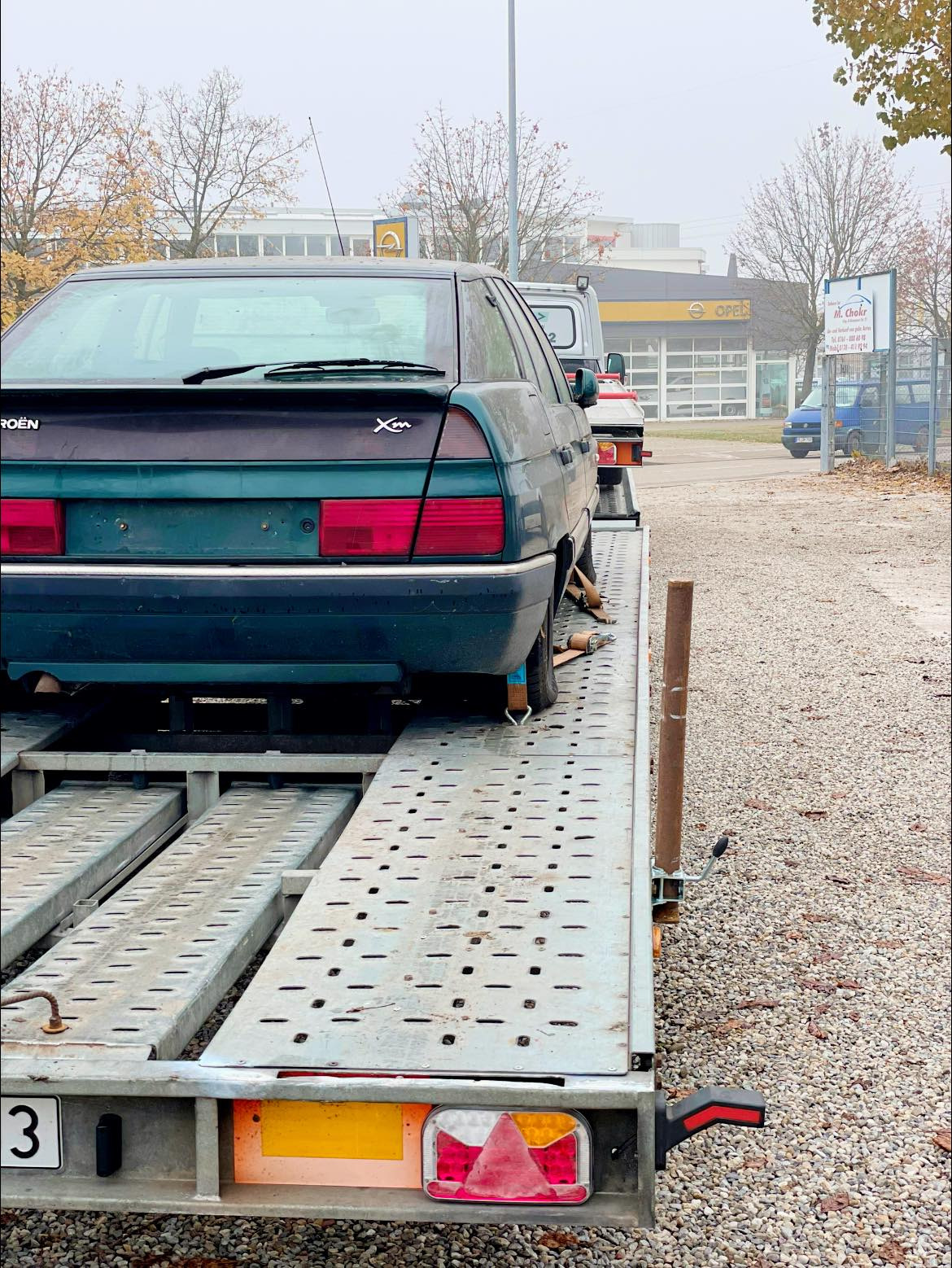 Zielony Citroen XM zabezpieczony pasami na metalowej platformie lawety, widok z tyłu, na tle zabudowań z logo Opla.