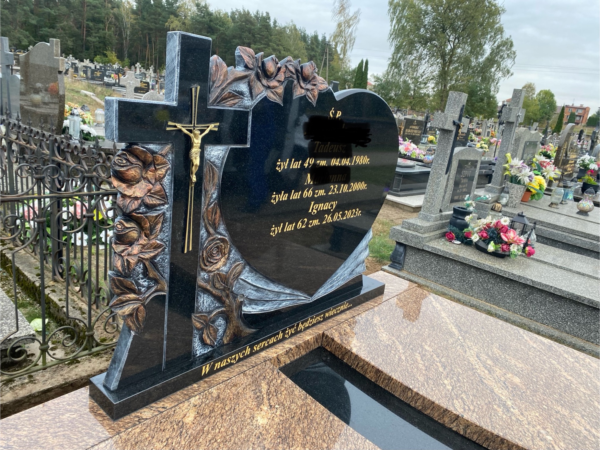 Czarny nagrobek z krzyżem i sercem, zdobiony motywem róż, na cmentarzu. Złoty krucyfiks i inskrypcje na płycie. Wyjątkowy projekt.