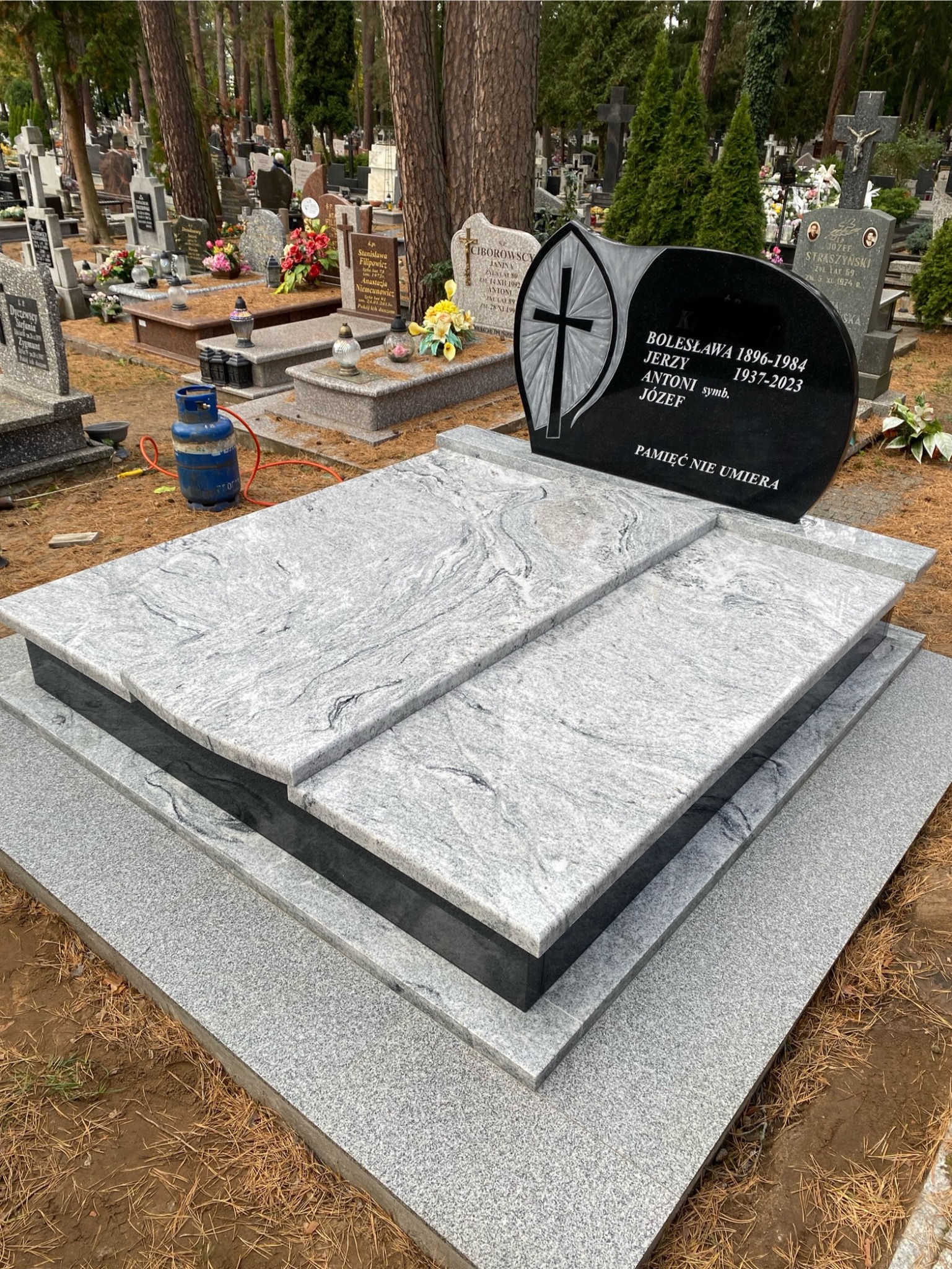 Szary nagrobek z granitu z czarną tablicą z inskrypcją na tle cmentarza. Widoczna butla gazowa. Kamienna konstrukcja nagrobka z wyraźnym wzorem.