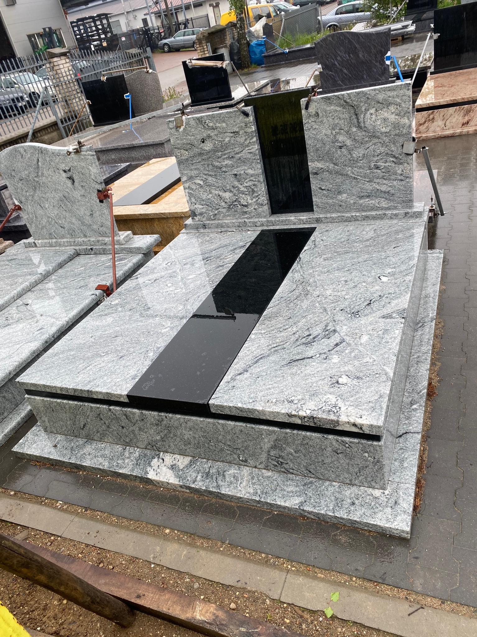 Szary nagrobek z czarnym pasem na płycie, ustawiony na zewnątrz w deszczowy dzień. Widoczne inne nagrobki w tle, co sugeruje ekspozycję w zakładzie kamieniarskim.