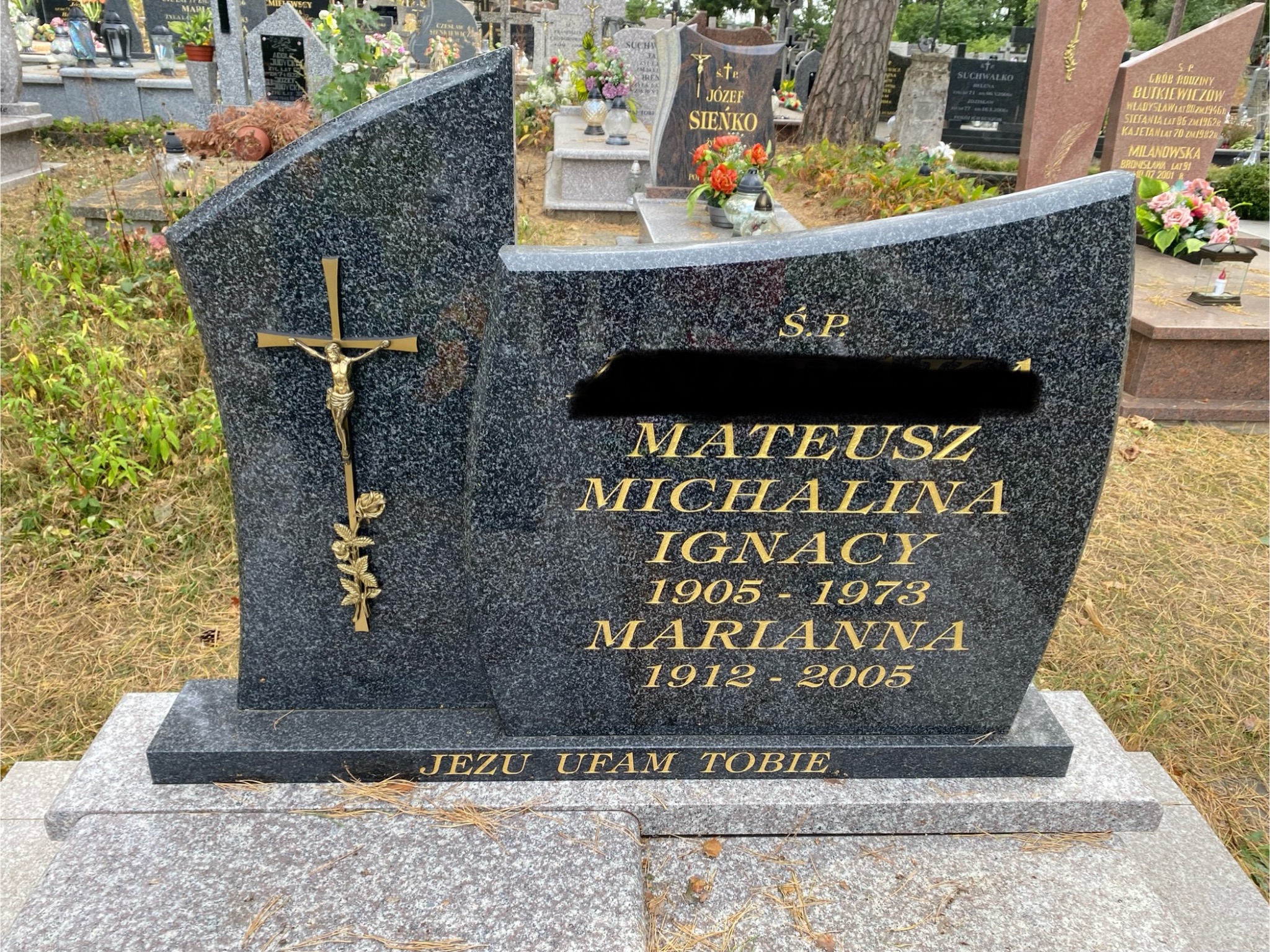 Granitowy nagrobek z krzyżem i inskrypcjami w złotym kolorze, otoczony innymi nagrobkami na cmentarzu. Widoczne napisy z imionami i datami.
