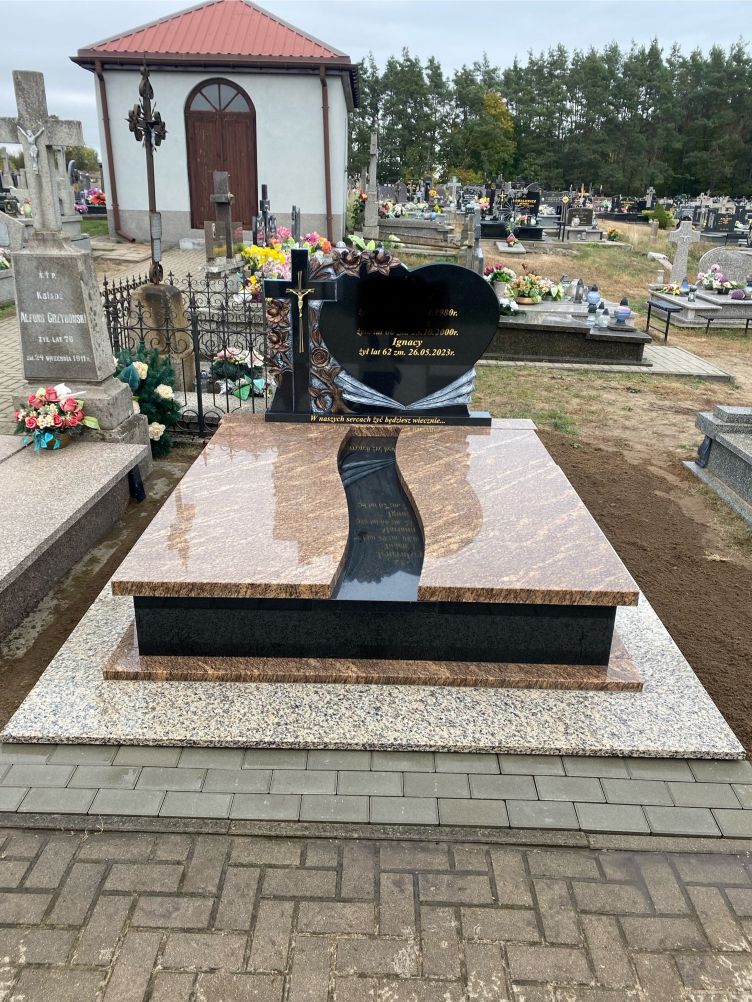 Granitowy nagrobek z czarnym sercem i złotym krzyżem, otoczony innymi nagrobkami na cmentarzu. Jasnobrązowy kamień z ciemnym, esowatym wycięciem.