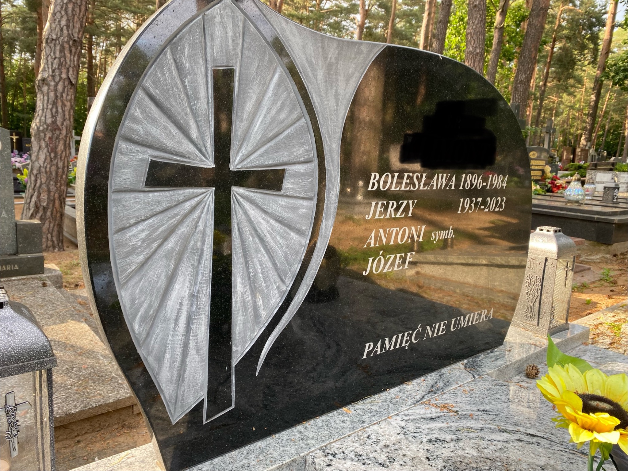 Czarny, granitowy nagrobek z delikatnym wzorem krzyża i wygrawerowanymi danymi, umieszczony na cmentarzu z widocznymi drzewami w tle.