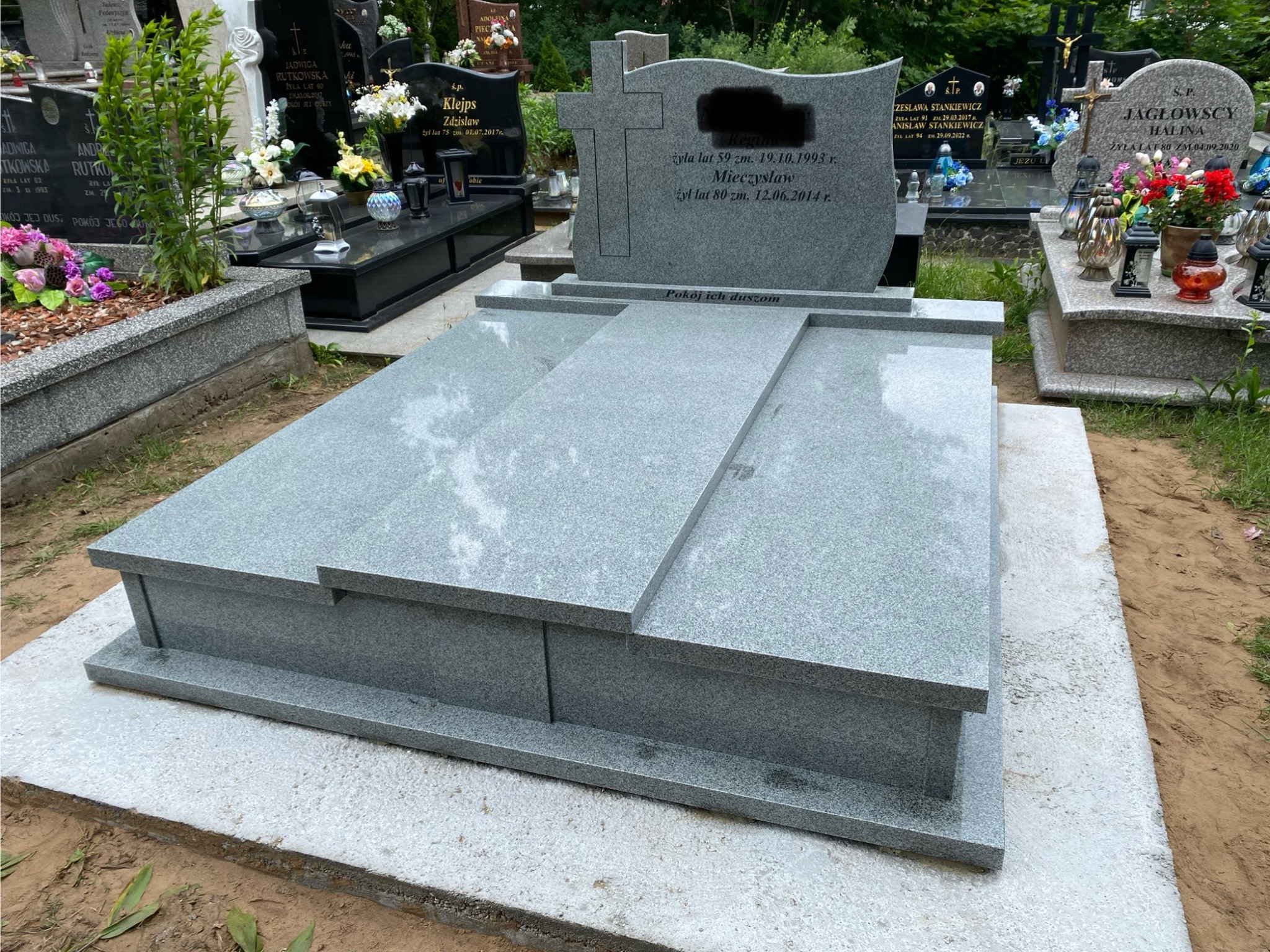 Szary, granitowy nagrobek z wygrawerowanym krzyżem i inskrypcjami na cmentarzu, otoczony innymi nagrobkami i zielenią. Widok z bliska, w dziennym świetle.