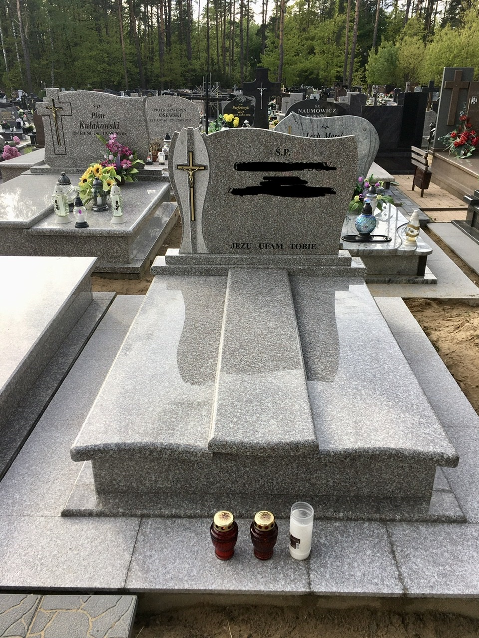 Nowy, jasnoszary pomnik z granitu z metalowym krzyżem po lewej stronie, ustawiony na cmentarzu w otoczeniu drzew, z trzema zniczami u podstawy.