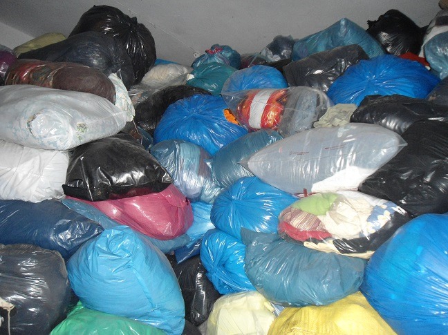 Stos worków z odzieżą używaną w różnych kolorach (niebieski, czarny, biały, różowy) pod sufitem magazynu. Część worków jest przezroczysta, ukazując zawartość.