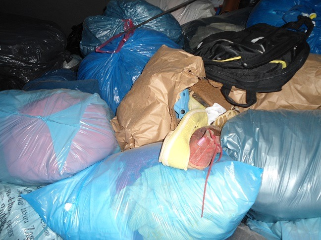 Stos worków z odzieżą używaną, widoczne buty i torba, różnorodne kolory i materiały. Ujęcie z bliska, ukazujące zawartość worków. Odzież sortowana.