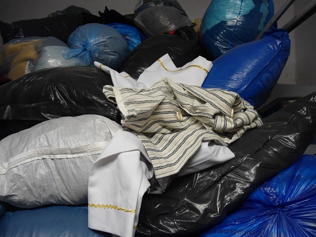 Stos worków z odzieżą używaną, widoczne ubrania z paskami i biała koszula z ozdobnym obszyciem, tworzące chaotyczny, ale uporządkowany wizualnie stos.