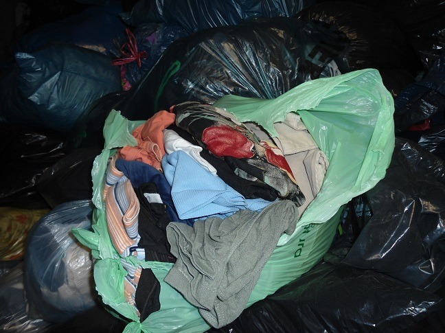 Stos worków z odzieżą używaną, częściowo otwartych, ukazujących różnorodne ubrania wewnątrz. Worki w kolorach czarnym, zielonym i niebieskim.