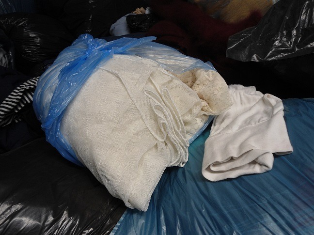 Worek z jasną odzieżą używaną, częściowo wystającą z worka, na tle innych worków z ubraniami. Widoczne faktury materiałów: dzianina, koronka.