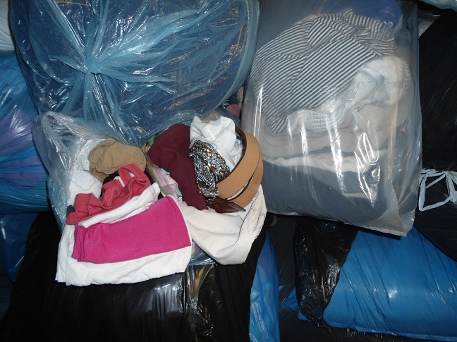 Wielkie worki z odzieżą używaną, niektóre przezroczyste, ukazują zawartość: paski, ubrania w paski, koszulki. Worki czarne i niebieskie w tle.