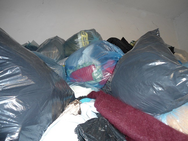 Stos worków z odzieżą używaną, wypełnionych ubraniami w różnych kolorach, w tym czerwonym swetrem, pod białym sufitem.