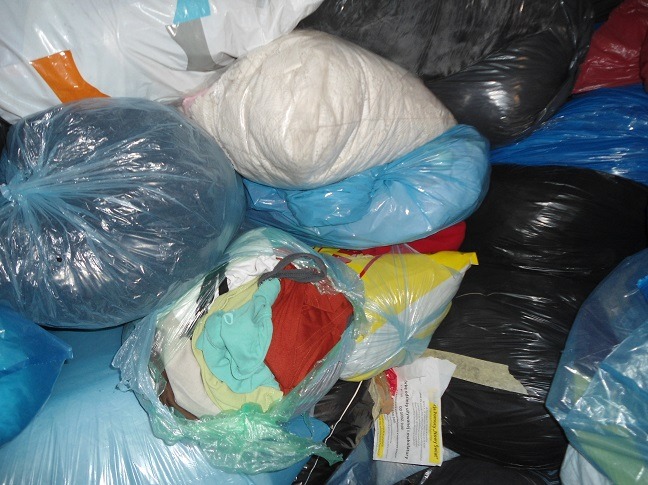 Stos worków z odzieżą używaną w różnych kolorach (czarny, niebieski, biały, żółty) i z różną zawartością, widoczne pojedyncze ubrania.