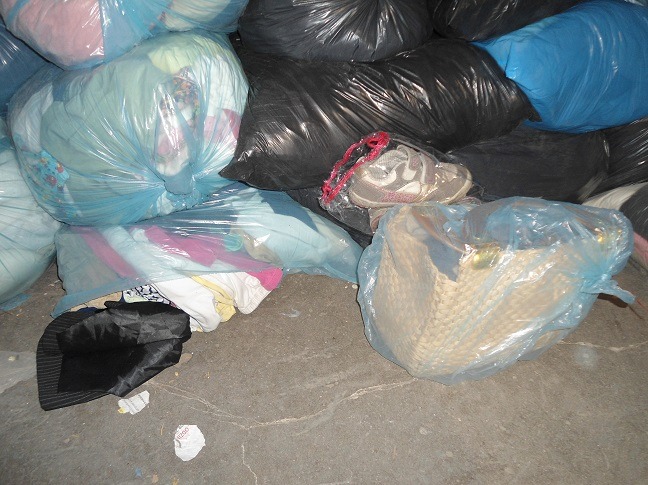 Stos worków z odzieżą i butami, niektóre w przezroczystych, niebieskich workach, inne w czarnych, na szarym betonowym podłożu.