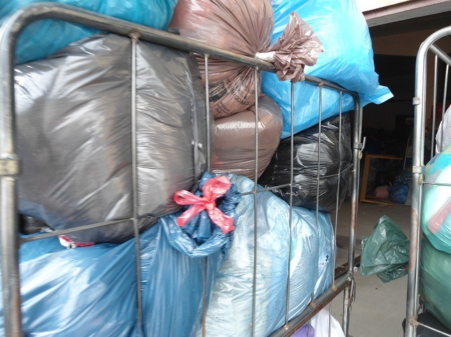 Wózek wypełniony workami z odzieżą używaną w różnych kolorach, z przewagą niebieskiego i czarnego. Widoczne metalowe elementy konstrukcji wózka.