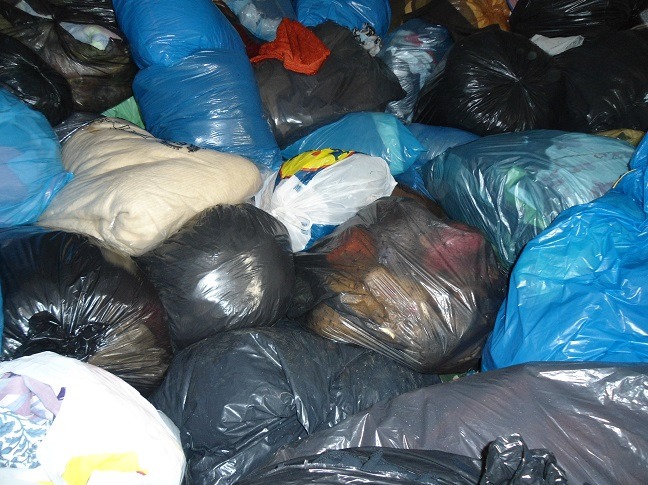 Stos worków z odzieżą używaną w różnych kolorach: czarnym, niebieskim i białym. Niektóre worki są przezroczyste, ukazując zawartość.