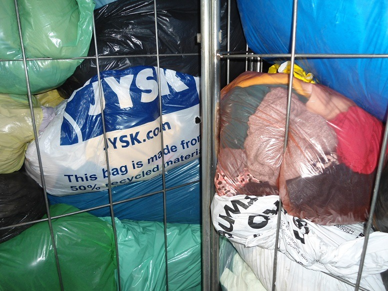 Wielobarwne worki z odzieżą używaną w metalowej klatce, jeden z worków z logo JYSK, widoczne różne kolory i faktury materiałów.